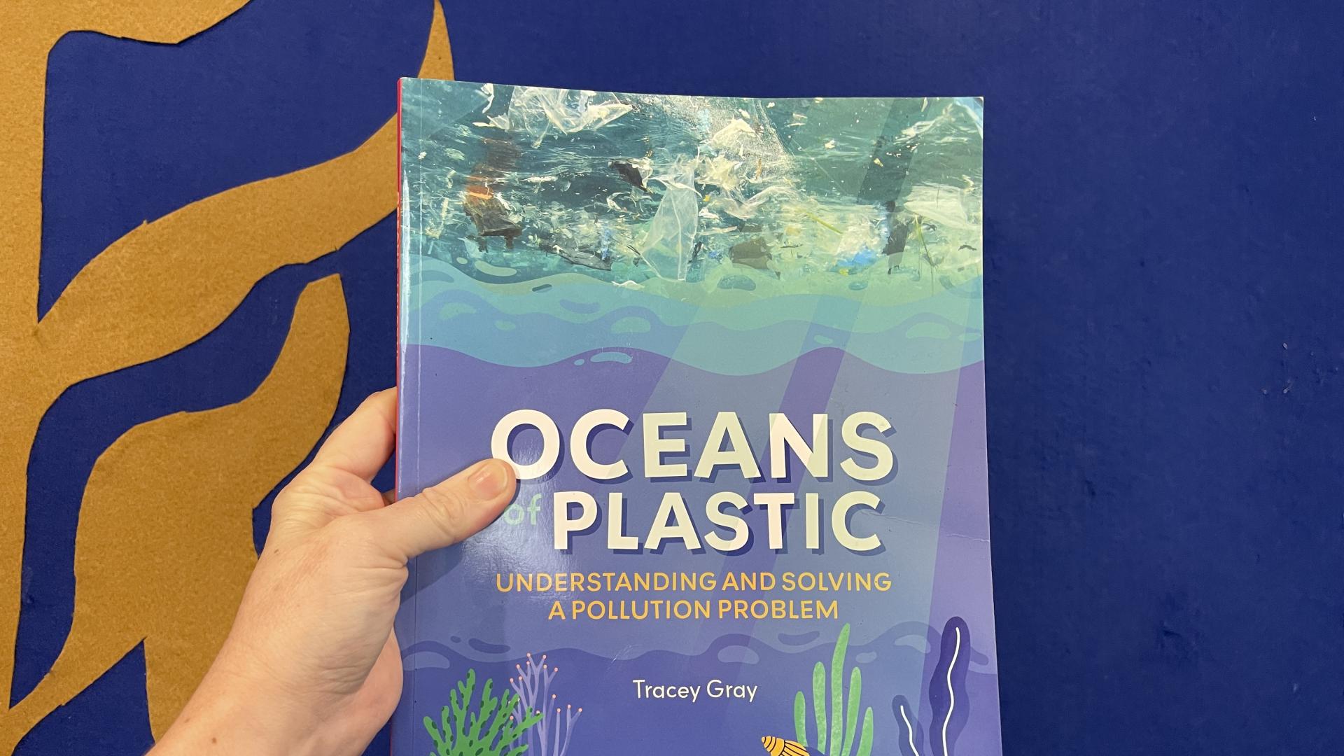 Oceans of Plastic. Understanding and solving a pollution problem book is held in front of a blue  wall with brown seaweed shapes on ited 